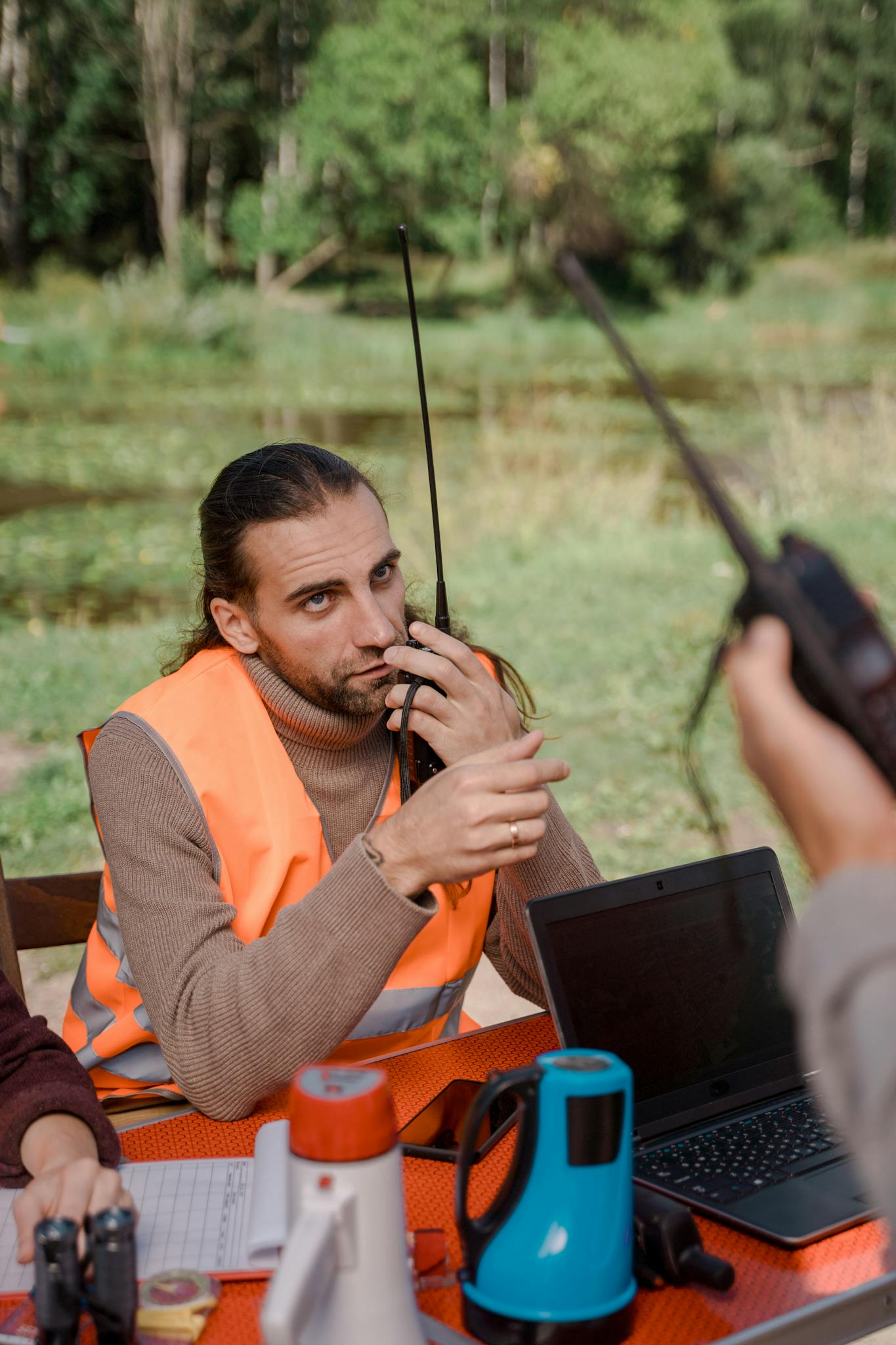 Man in orange vest using walkie talkie at outdoor search operation with laptop.